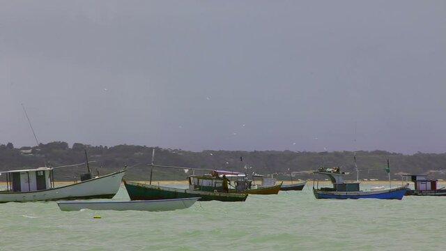 Artisanal fishing boat sailing among other anchored boats, Coroa Vermelha beach, sea with ripples, sky with clouds