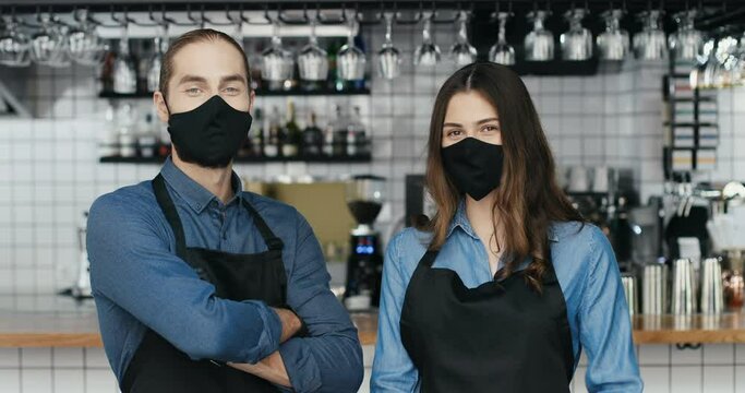 Portrait young Caucasian cheerful couple of bartenders in masks and gloves looking at each other, smiling to camera happily. Happy waiter and waitress in cafe. Joyful man and woman, baristas at work.