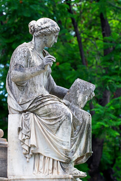 Marble statue on the base of the statue of Ioannis Varvakis in the garden of the Zappeion Palace of Athens, with trees blurred in the backround
