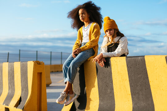 Fashionable multiethnic children sitting on street barrier fence