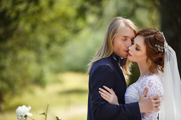 Fototapeta premium Young Bride and Groom couple in a summer garden. Tender holding each other. Redhead woman and blonde man with long hairs. Young family outdoor in nature. Love and tenderness