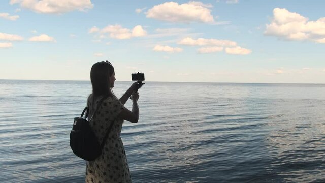 Back view portrait of woman making video of beautiful seascape using smartphone and monopod at sunset