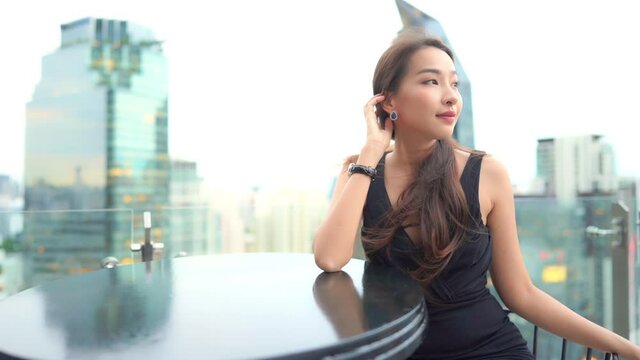 While sitting at a rooftop bar the wind tosses the hair of a beautiful young woman in a black cocktail dress.