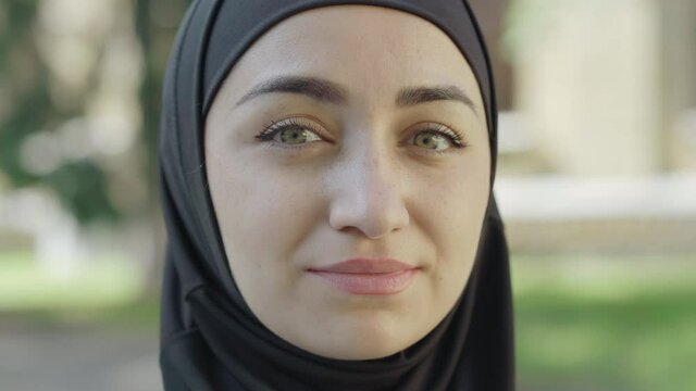 Close-up face of young beautiful woman in traditional hijab looking at camera and smiling. Portrait of confident charming Muslim lady posing outdoors on sunny day. Diversity and beauty concept.