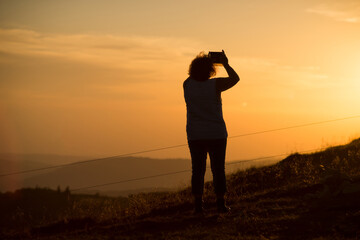 Portrait  on back view of woman silhouette standing at the top of the mountain taking a photography  with his smatphone in hands on sunset background