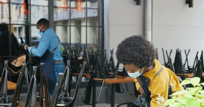 African American couple of baristas, male and female in masks and gloves cleaning desks and chairs in bar. Waiter and waitress clean and preparing public place in cafe. Reopen concept. Preparation.