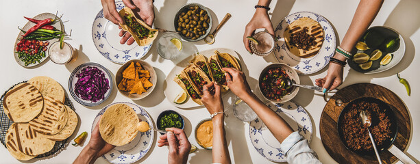Friends having Mexican Taco dinner. Flat-lay of beef tacos, tomato salsa, tortillas, beer, nachos and peoples hands over white table, top view. Mexican cuisine, gathering, feast, comfort food concept