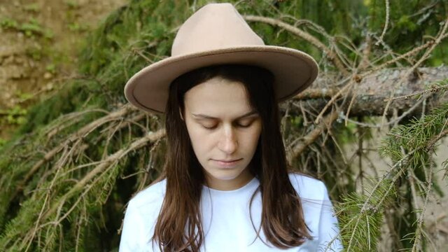 Close up portrait of young attractive caucasian hipster woman in hat standing near tree look straight at camera, dressed in white t-shirt. Explore travel real wilderness lifestyle. Slow motion