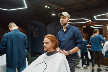 Young professional barber serving client, young redhead guy sitting in barbershop chair