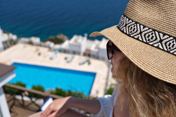 Beautiful young woman in straw hat on white terrace balcony of house or hotel looking to swimming pool and Sea View