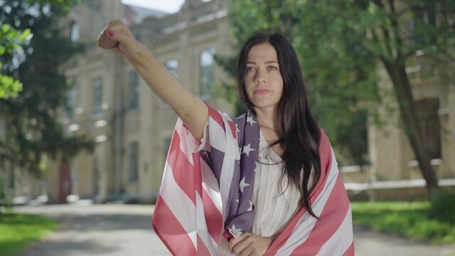 Portrait of confident young woman in American flag making liberty hand gesture at university yard. Serious beautiful brunette student posing for freedom at the background of college on sunny day.