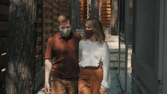 Tracking shot of young man and woman in face masks walking together outside of their vacation home surrounded by wooden fence