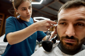 Close up shot of a professional barber girl or female hairdresser working with hair clipper, making trendy haircut for a handsome bearded man