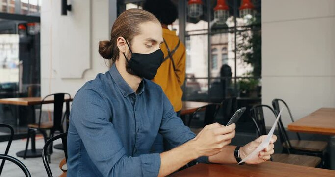 Caucasian man client in mask siting at table in cafe outdoor and scanning QR code with smartphone for reading menu. Technologies during covid-19 pandemic. Male customer in bar making order on phone.