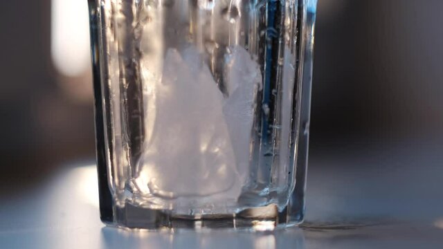 Wet empty transparent glass with ice pieces. Concept. Close up of putting ice pieces into the faceted glass standing on white table on blurred background.