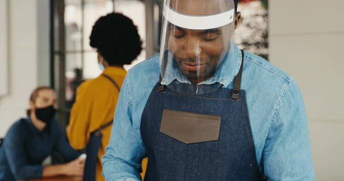 African American young handsome cheerful man waiter in plastic mask and gloves taking order with tablet device in cafe at terrace. Pandemic measures in public place. Male talking with client.