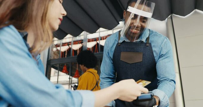 Caucasian female client in cafe outdoor terrace and African American male barrista in masks talking. Woman customer in restorant paying with credit card outdoors. Mixed-races. Multiethnic. Pandemic.