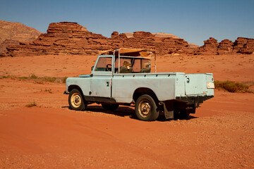 Fototapeta na wymiar A vintage turquoise colored pick up truck is abandoned on red sands of the Wadi Rum desert. This is a very old and rusty vehicle, poorly maintained and is broken down.