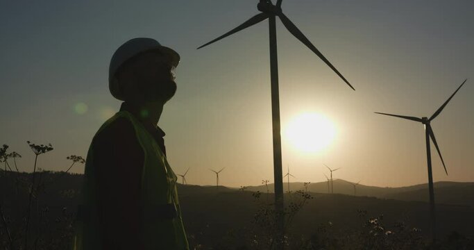 A young engineer enters the frame and then examines the windmill in front of him at sunset