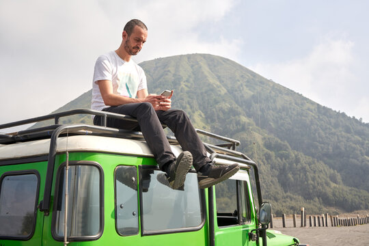Man visiting Bromo volcano by jeep and sitting on the top of the car using a mobile phone