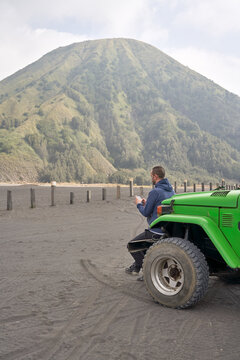 Man visiting Bromo volcano by jeep and sitting on the hood of the car using a mobile phone