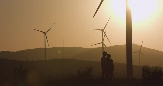 Two young men standing near the wind turbines pointing their hand forward. Silhouette shooting. Sunset