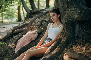 girls brunette in the evening light sits under a tree in the roots in a white vest and shorts