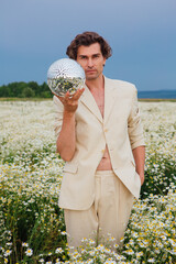 Tall handsome man standing in camomile flowers field holding mirror disco ball.