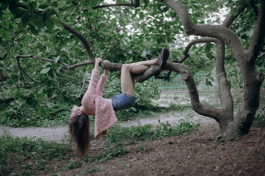 a girl with long black hair in a pink soft jacket hangs on tree branches like a monkey upside down
