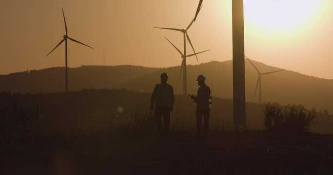 Two engineers walk towards the camera turning on the windmill. Sunset