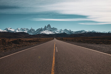 Panoramic image of a long straight asphalt road leading to frozen snowy mountains in the horizon and some clouds in the blue sky. Patagonia, Chile.
