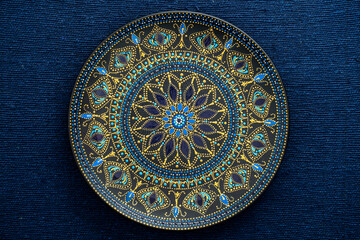 Decorative ceramic plate with black, blue and golden colors, painted plate on background of blue fabric, dot painting