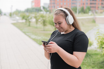 A beautiful fat girl in black sportswear listens to music on headphones and text messages on the...