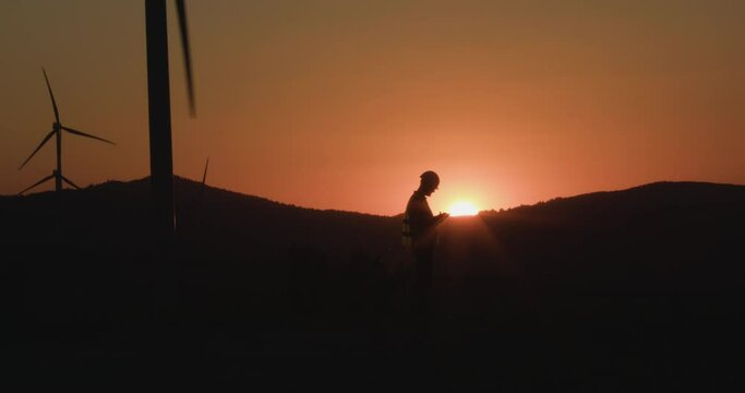 Engineer writes information in a tablet looking at windmills at sunset. Silhouette shooting. The concept of environmentally friendly energy