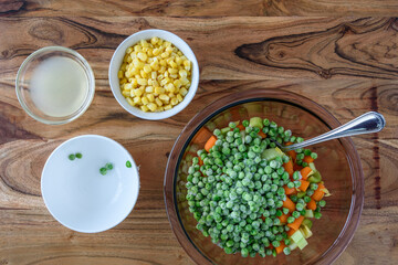 Making a cold salad, glass bowl with chopped celery, carrots, and frozen peas, bowls with frozen corn, and fresh squeezed lime juice on wood cutting board
