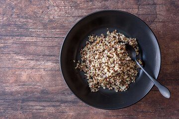 Cooked tri-color quinoa in a ceramic black bowl, with a black spoon, on a rustic wood background
