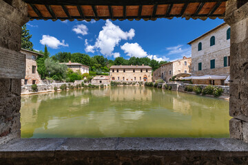 An ancient medieval termal pool framed by a wooden canopy, under a blue summer sky with puffy clouds