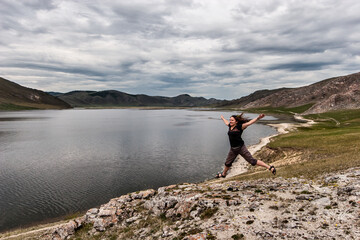 Fototapeta na wymiar A young girl laughs and joyfully jumps on a rock near the lake. Beautiful mountains in the background. Freedom and joy. The sky is overcast. Horizontal.