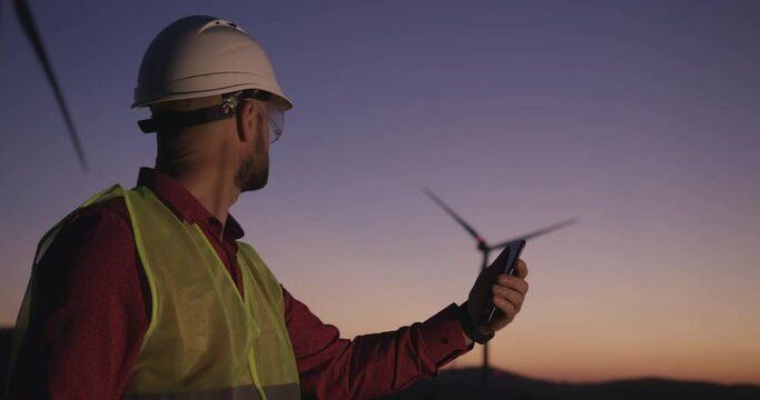 Engineer browses information on the phone standing on the background of windmills looking away to the left at sunset