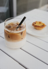 Glass with ice latte and black straw on white table. Dessert in the background. World coffee day.