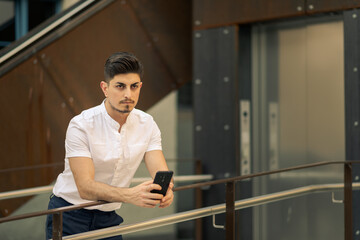 Urban portrait of young arabic man in business outfit holding his mobile phone while looking...