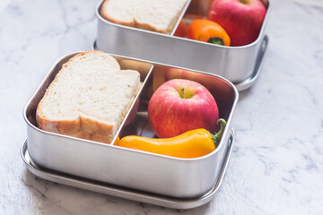 Close up view of stainless steel lunch containers with healthy food, including wholemeal sandwich, apple, carrots and bell pepper on grey marble background (selective focus) - Powered by Adobe