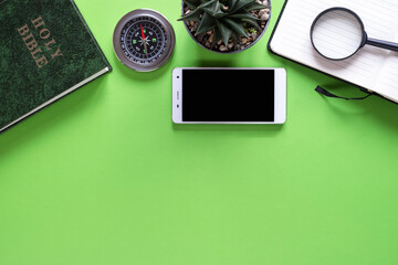 Top view of Holy Bible, phone, plant, compass, magnifying glass, and notebook on green background