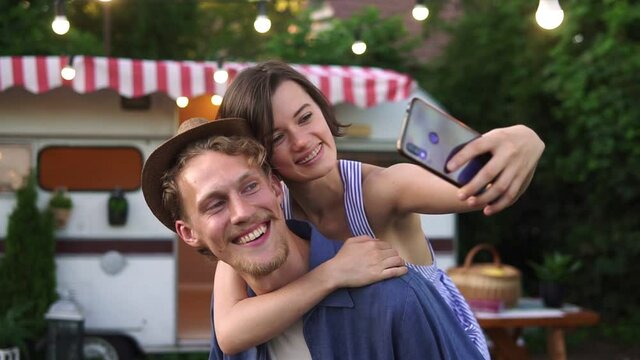 Happy young lovely couple having fun near trailer in the park, man piggybacking girl. Smiling girl in summer dress and hat doing selfie using smartphone. Perfect picture of cheerful, young couple on