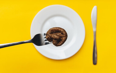 Artificial cat feces lie on a fork close-up. White plate and a knife with a yellow tablecloth. The concept of fake news, inept cook, crappy restaurant and bad food. Smelling joke, taste of shit.