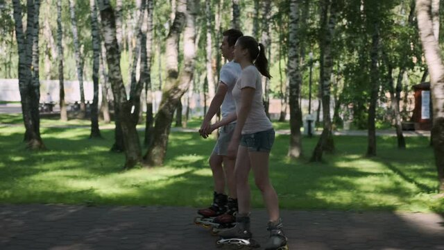 Happy young couple of teenagers rollerblading in park holding hands.