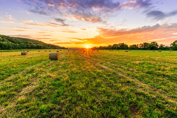 Scenic view at beautiful sunset in a green shiny field with hay stacks, bright cloudy sky , trees and golden sun rays with glow, summer valley landscape