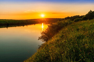 Fototapeta na wymiar Scenic view at beautiful spring sunset with reflection on a shiny lake with green reeds, bushes, grass, golden sun rays, calm water , sky and glow on a background, spring evening landscape