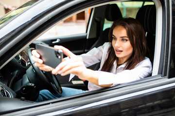 Happy young woman holding mobile phone and taking photos while driving a car. Smiling girl taking selfie picture with smart phone camera outdoors in car. Holidays and tourism concept