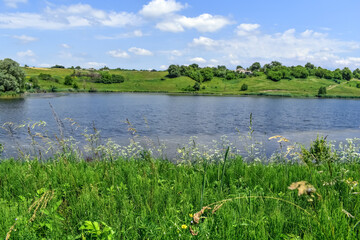 Lake in the middle of meadows with green lush grass on a sunny spring-summer day. Beautiful rural landscape with a pond. Panorama of the village of Uhroidy (Ugroedy) in the Sumy region (Ukraine)